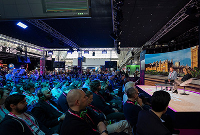 Photo: a conference space with rows of chairs filled with people on the left half of the image, looking towards the right of the image where two people are sat in chairs on a stage.