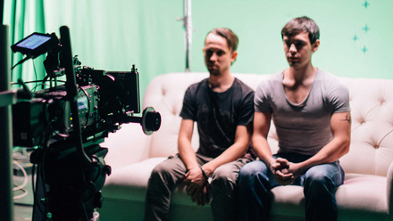Two actors sit on a sofa with a green screen behind them, facing directly into the lens of a camera in the left-foreground of the image.