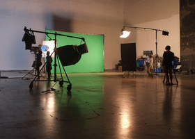 Photo of a large studio with a green screen and lights set up within it and a couple of crew members.